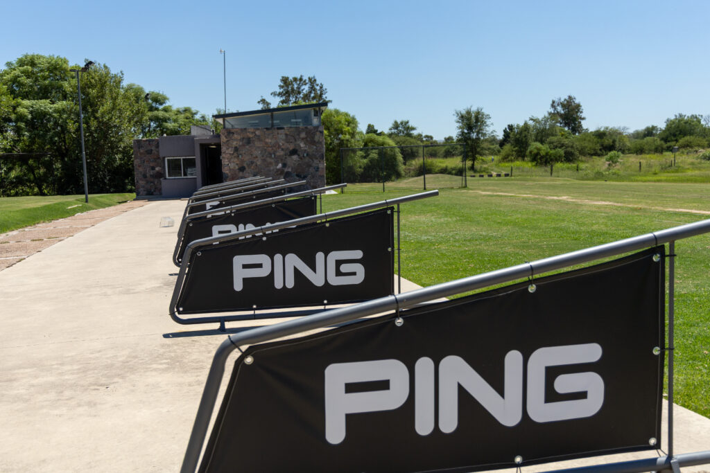 Vista general del driving con la casilla de atención al fondo y los tees techados con cartelería de PING.